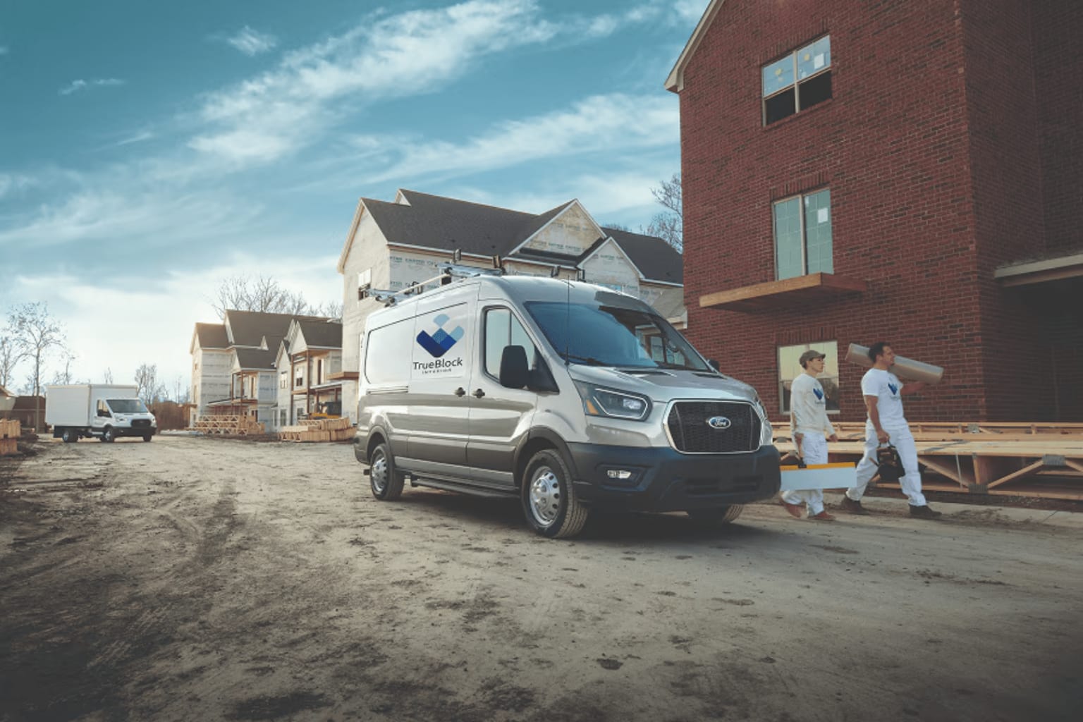 A silver van is parked on a dirt road in front of a brick house under construction, with two workers carrying lumber and other houses in the background.