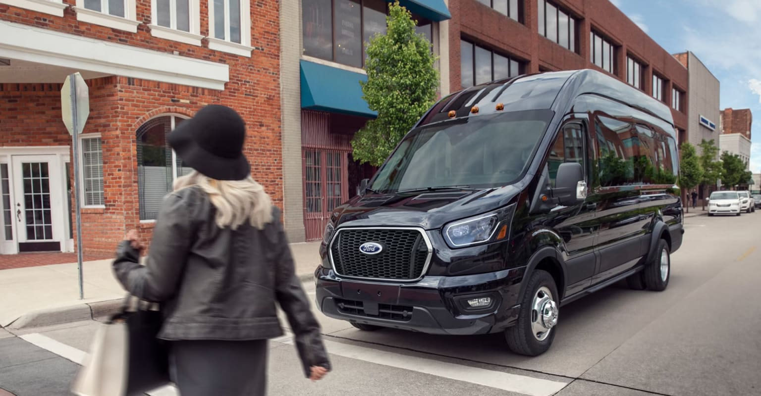 A person in a gray coat walks past a large black van parked on the street, with brick buildings and storefronts in the background.