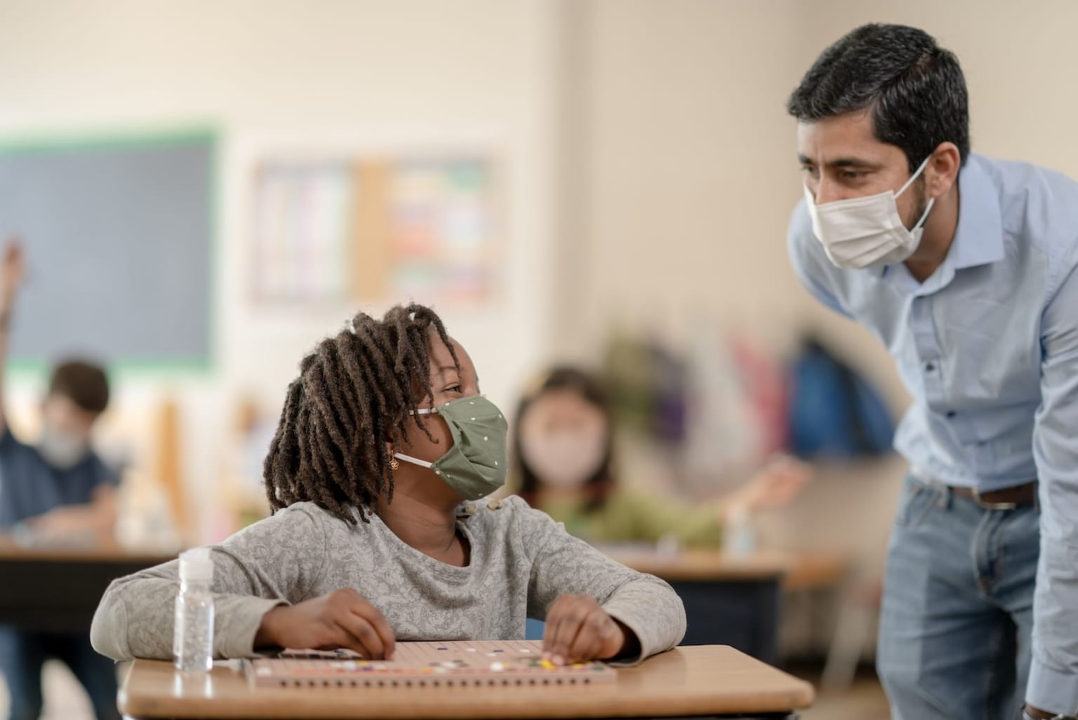 a man wearing a face mask standing next to a young girl in a classroom with other children in the background