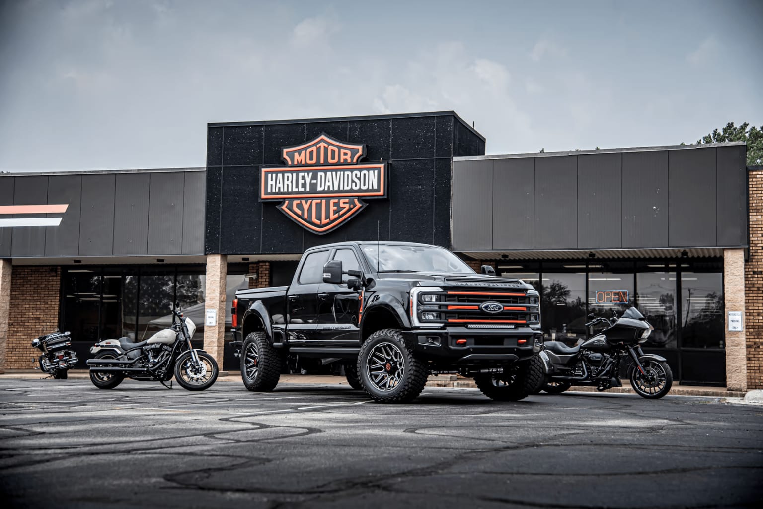 A large black pickup truck and two motorcycles parked in front of a dealership building with the company's iconic logo displayed.