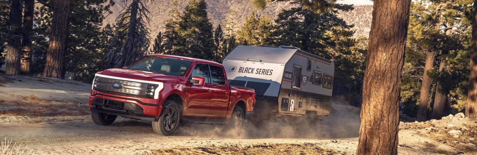 A red pickup truck towing a large camper trailer driving down a dirt road surrounded by tall pine trees and a rocky mountainous landscape.