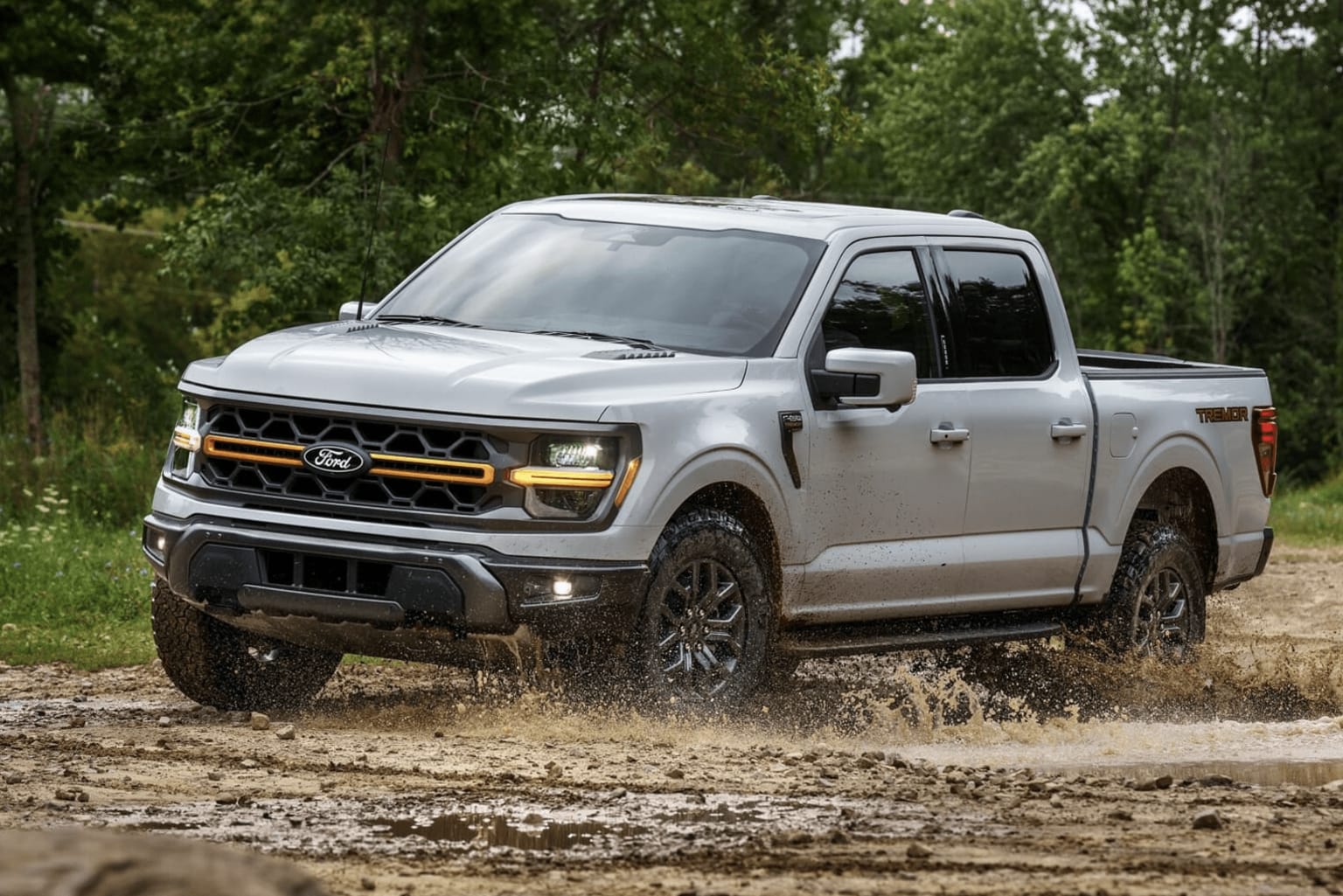 A silver pickup truck is parked on a dirt road surrounded by lush green foliage, kicking up dust as it navigates the rugged terrain.