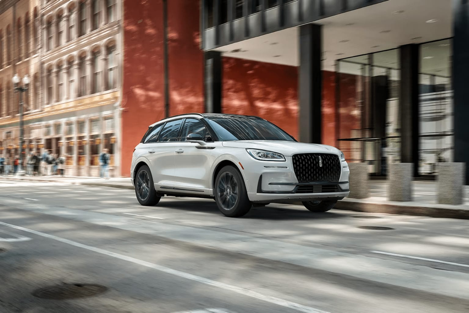 A white luxury SUV driving down a city street with tall brick buildings and pedestrians visible in the background.