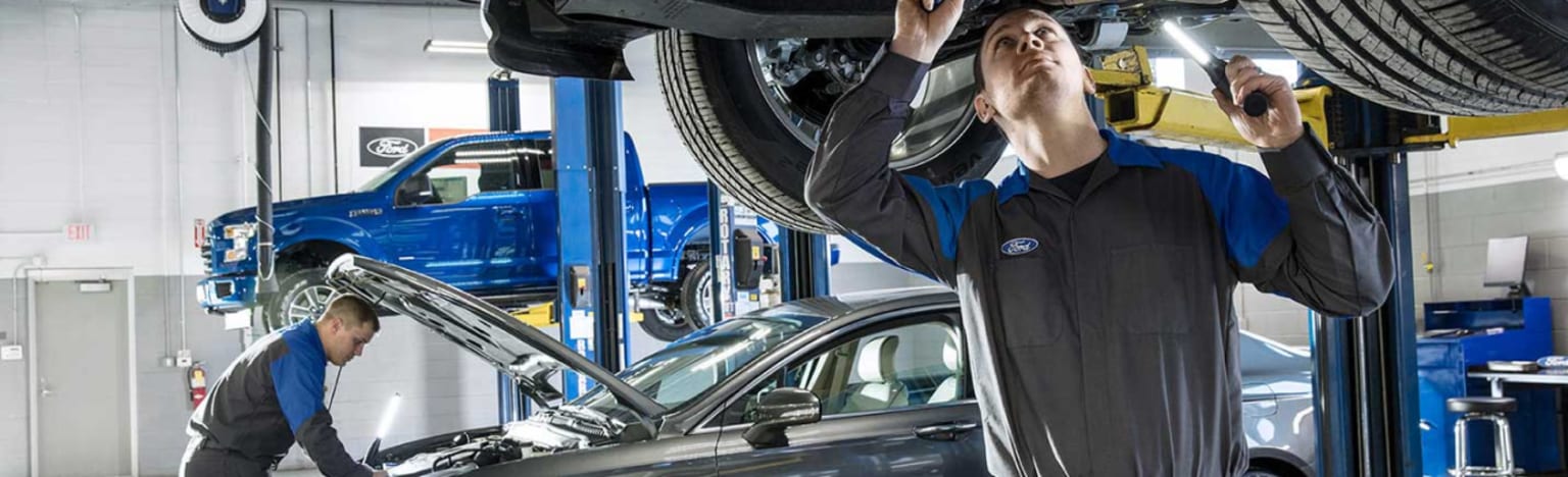 Two mechanics working on cars in a brightly lit garage, with a blue truck lifted in the background.