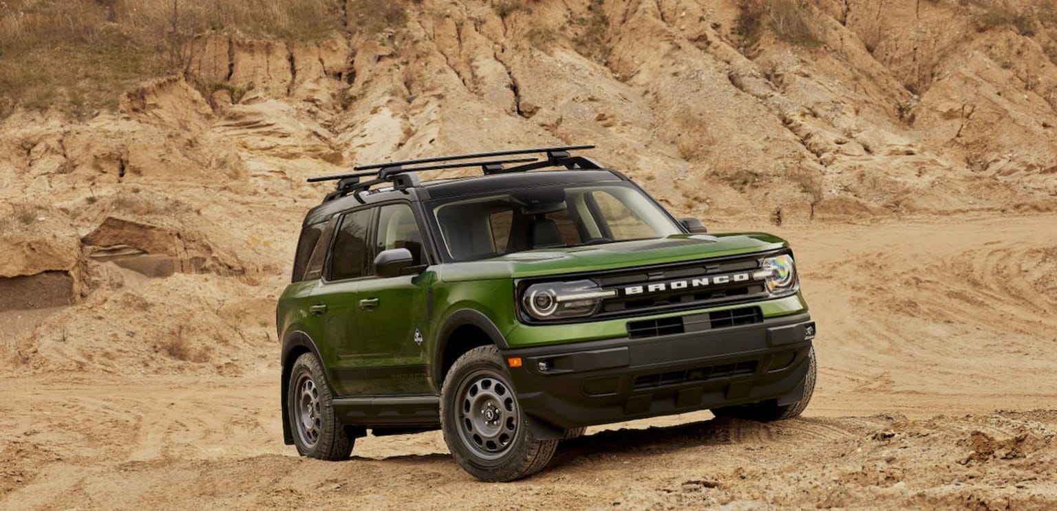 a green truck driving on a dirt road near a rocky hill side with a hill in the background and a cliff in the foreground