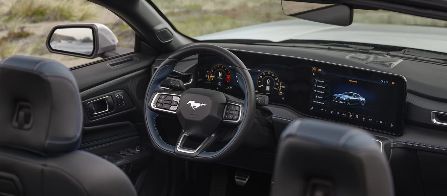 Interior of a modern vehicle with a steering wheel, dashboard, and various controls and displays. Background shows a natural outdoor setting, suggesting the vehicle is being driven in a rural or remote area.