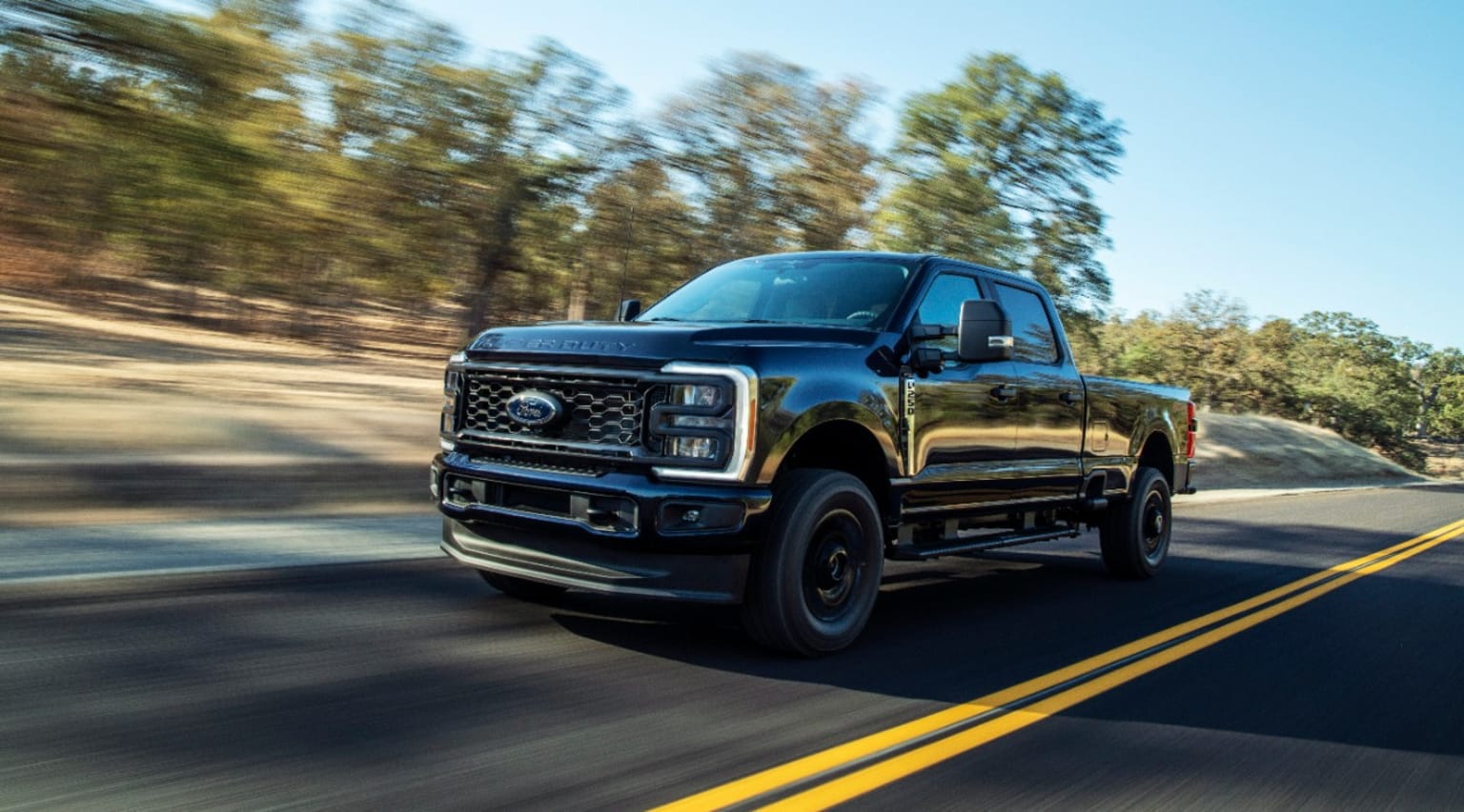 a black truck driving down a road next to a wooded area in the day time with a blurry image of the front end of the truck