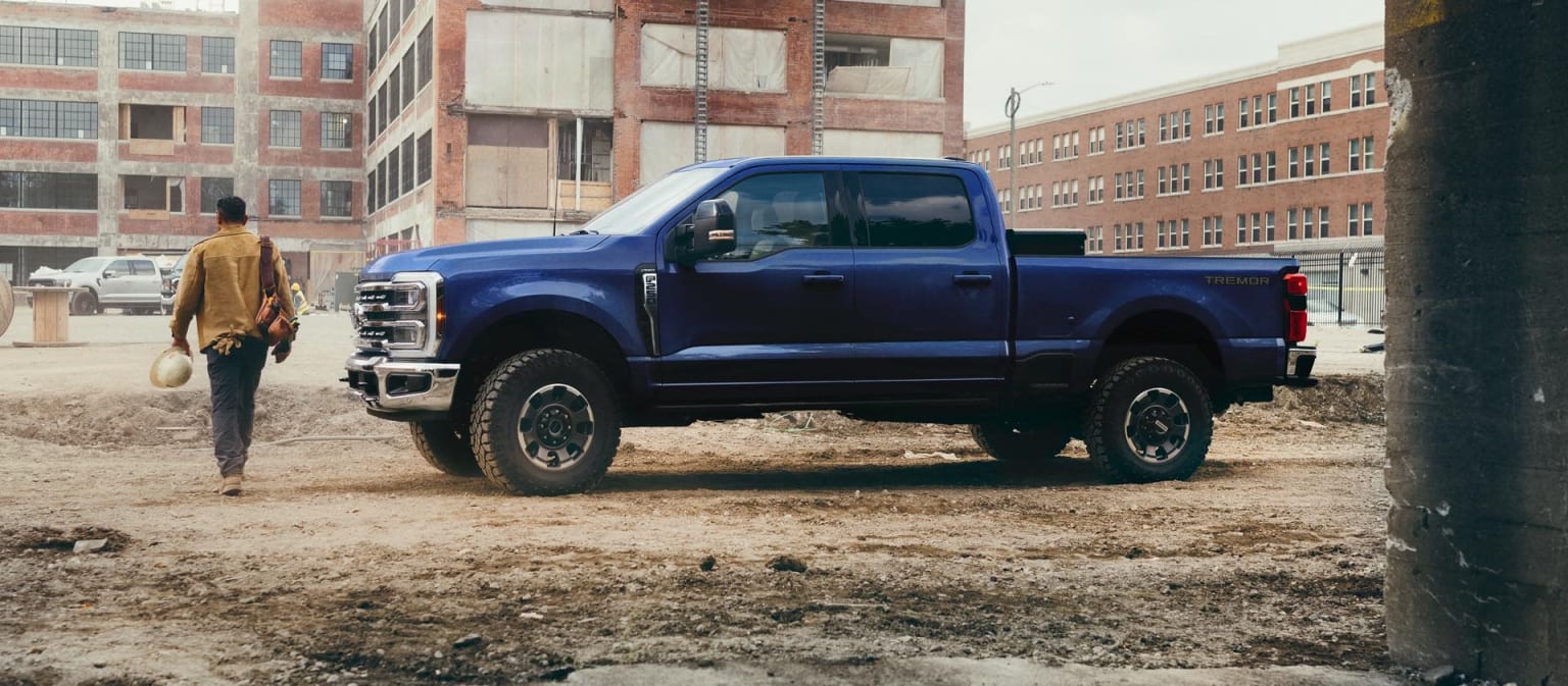 A large, blue pickup truck with oversized tires on a muddy construction site, with a person in a yellow jacket walking in the background.