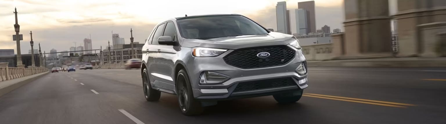 A silver SUV with a bold front grille and sleek design driving on a city street, with tall buildings and a cloudy sky visible in the background.