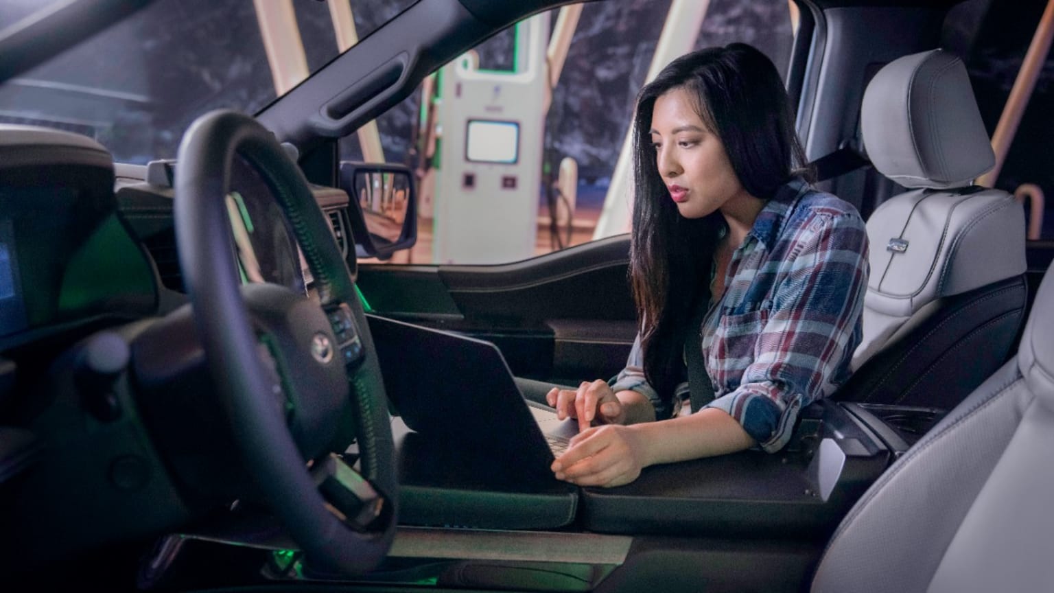 a woman sitting in the driver's seat of a car using a laptop computer while holding a cell phone