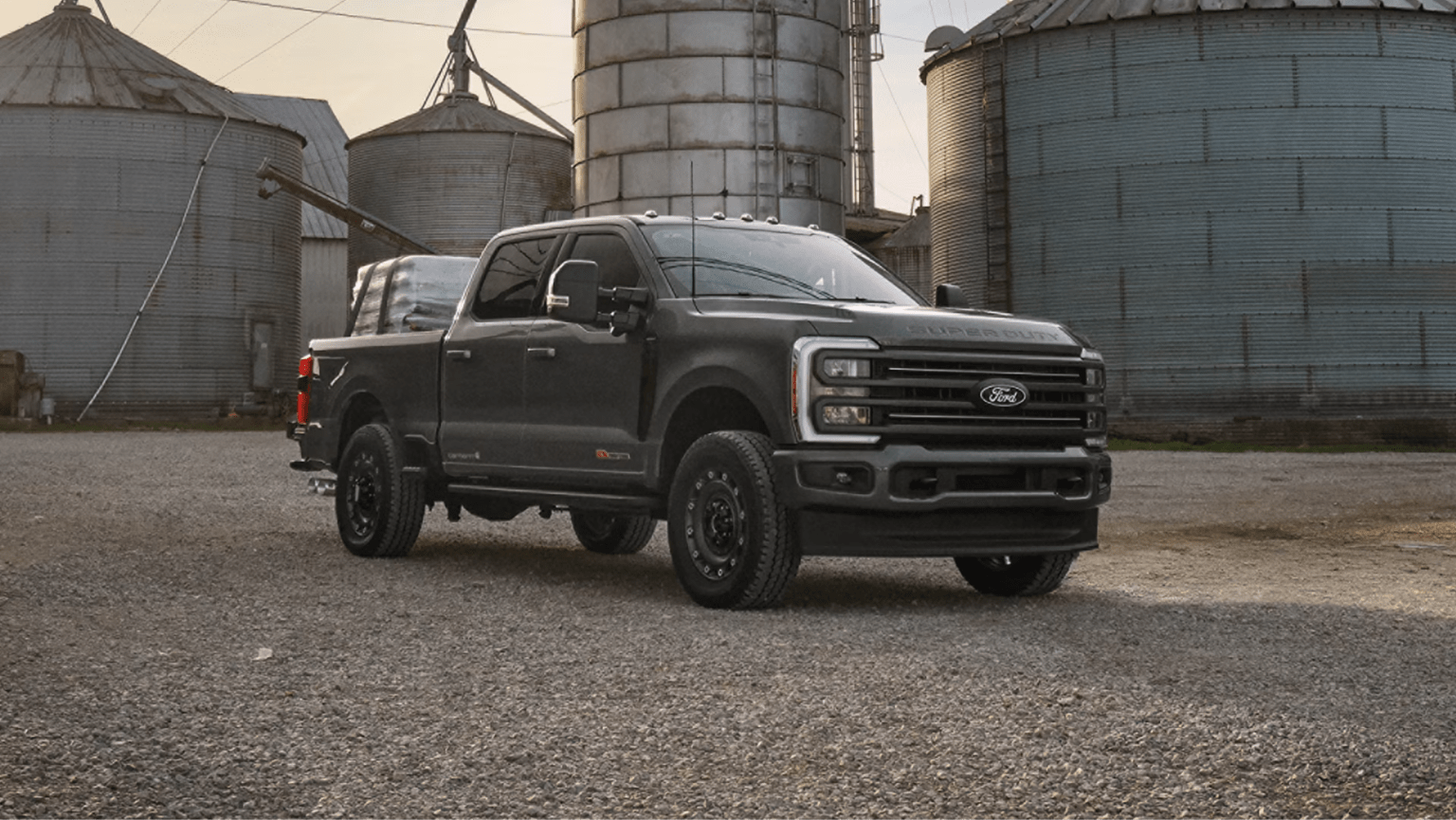 A dark grey pickup truck parked on a gravel lot in front of metal grain silos.