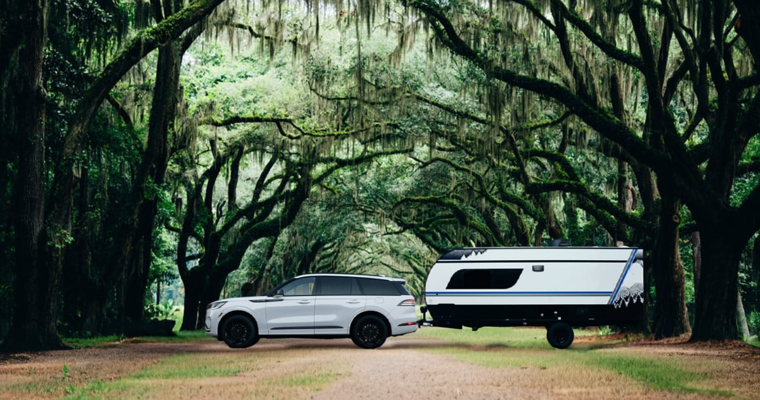 A white SUV towing a camper trailer on a winding dirt road surrounded by a lush, green forest canopy.