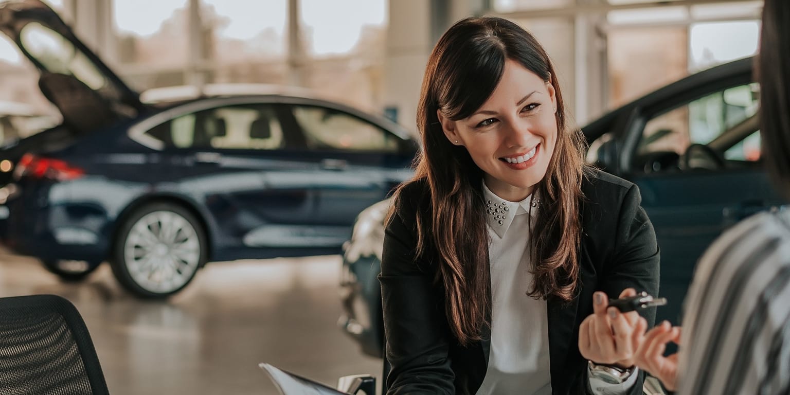 A young woman with long brown hair is smiling and standing in front of several cars in what appears to be an automobile showroom or dealership.