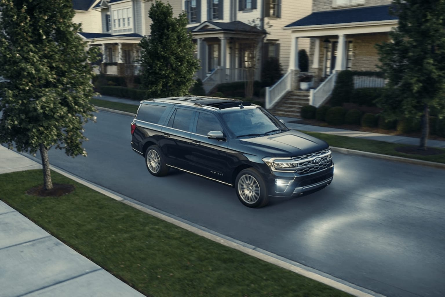 The image displays a large black SUV driving down a residential street with well-maintained houses and lush greenery in the background.