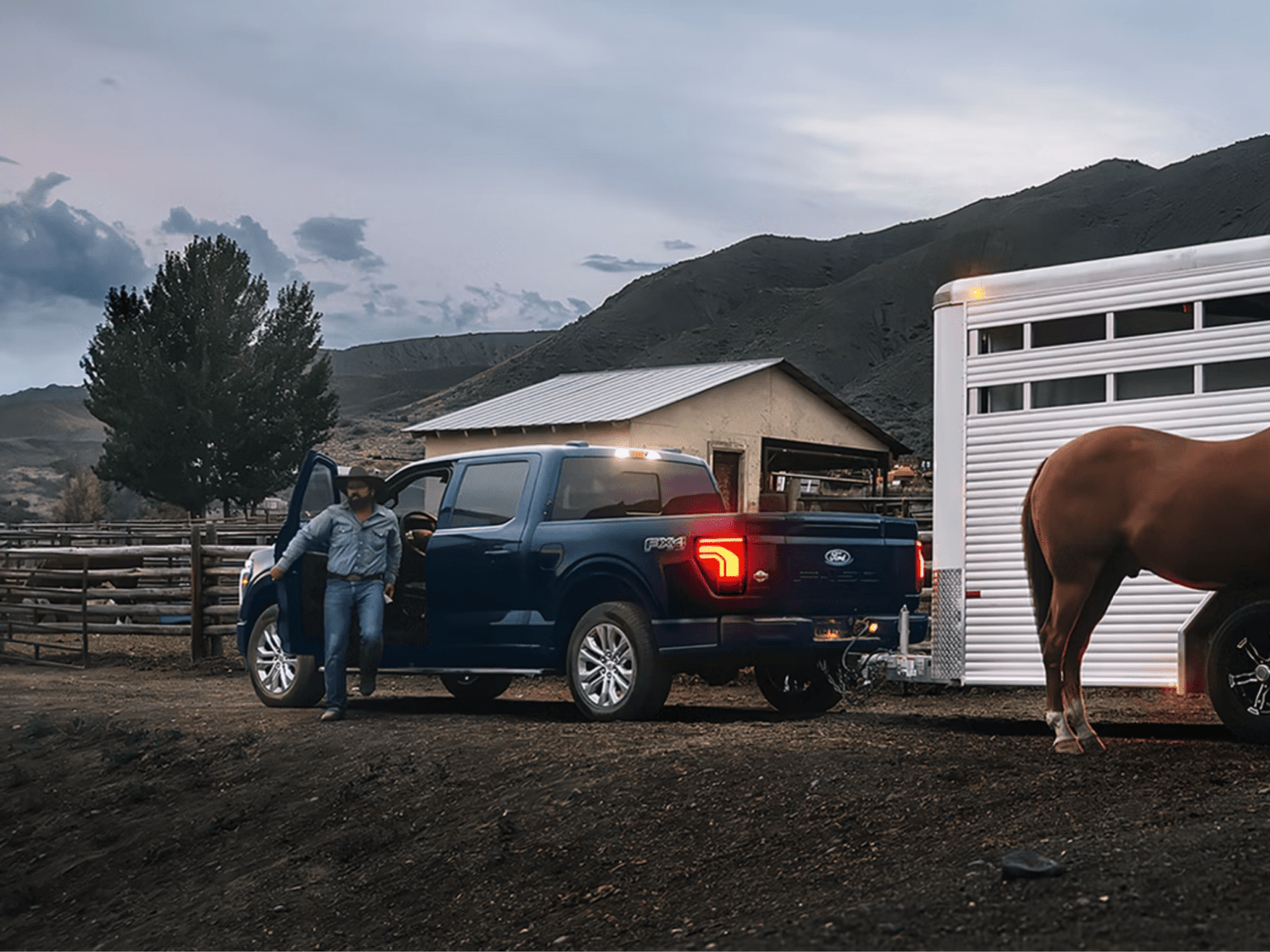 A cowboy standing by a dark blue pickup truck hitched to a horse trailer, with a barn and rolling hills in the background.