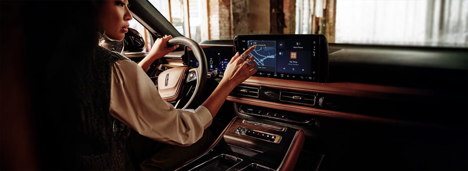 A person driving a car with hands on the steering wheel, looking at the navigation display on the dashboard.