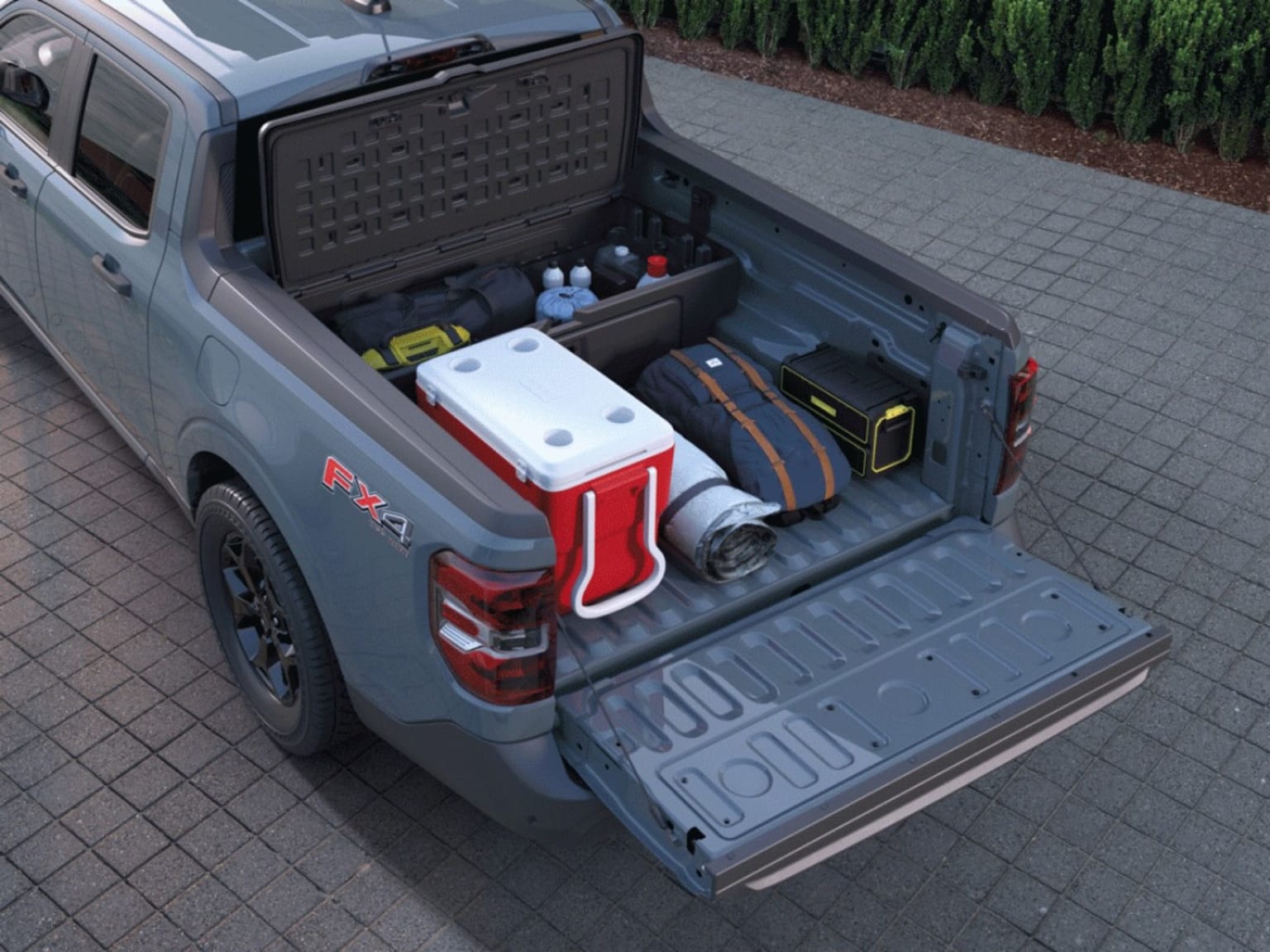 A grey pickup truck with a loaded bed on a cobblestone driveway in front of a green hedge.