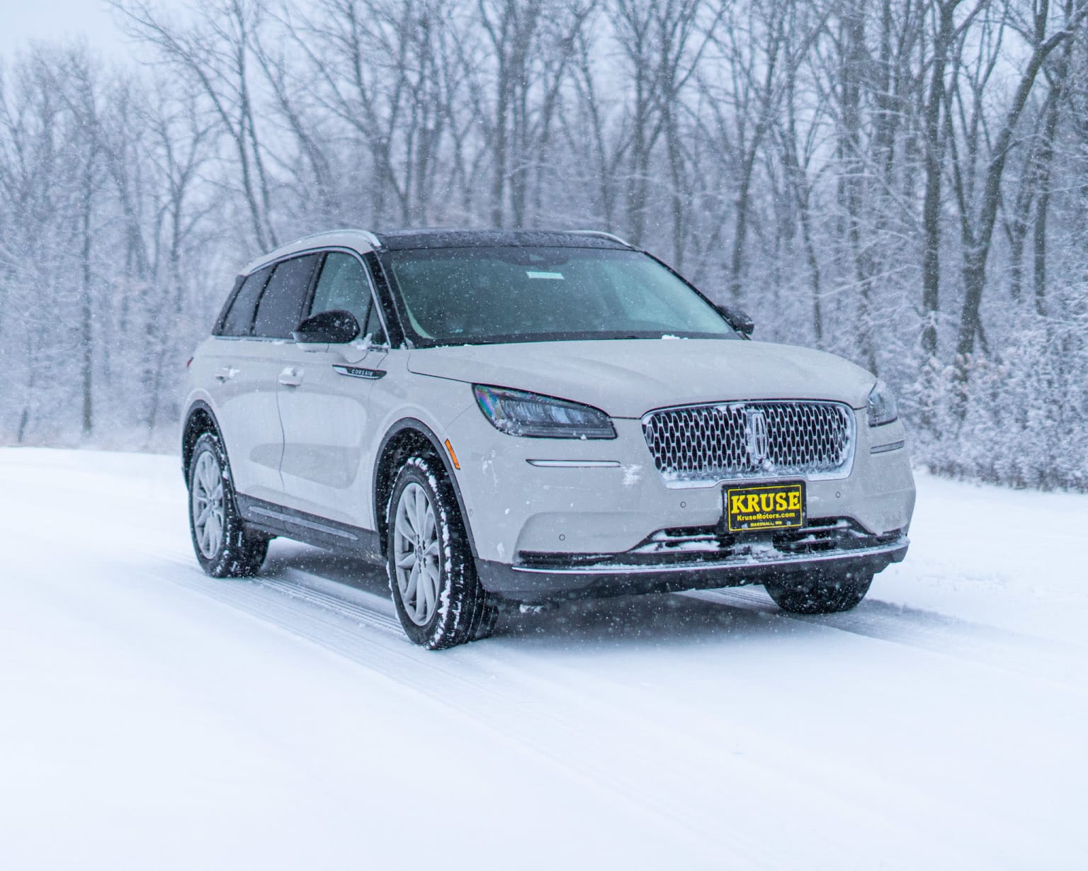 a white suv driving down a snow covered road in the woods with trees in the background and a yellow license plate on the front of the vehicle