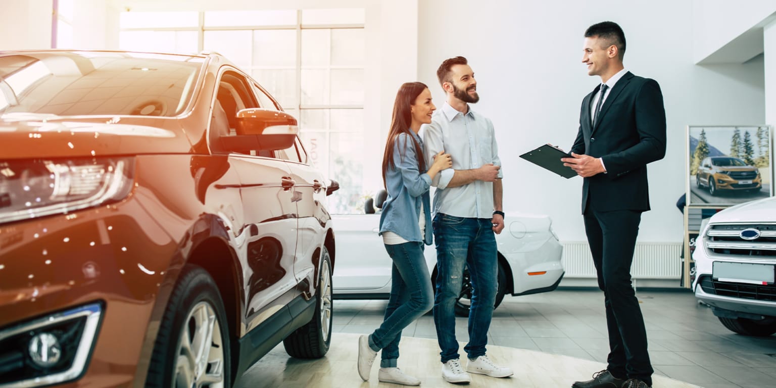 A couple talks with a salesman in a brightly lit car dealership, with a red SUV in the foreground and other cars and advertisements in the background.