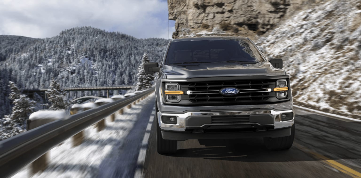 A rugged pickup truck navigates a snowy mountain road, with a backdrop of snow-capped peaks and a forested landscape.