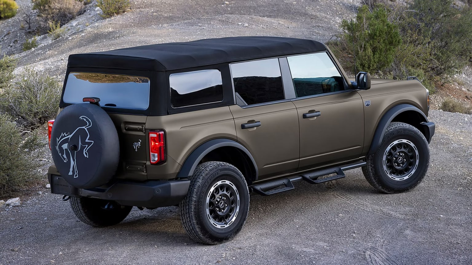 A bronze SUV with a black soft top is parked on a dirt road with a rocky, arid landscape in the background.