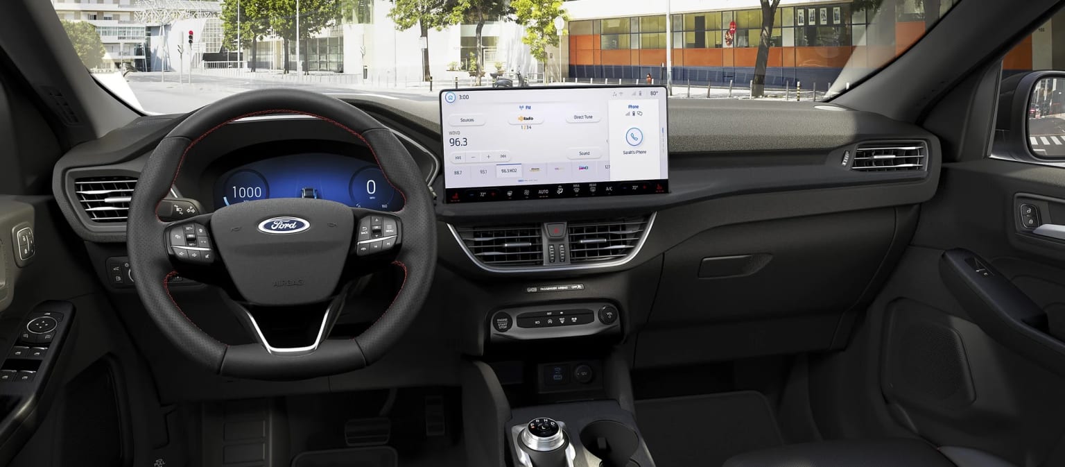 The image shows the interior of a vehicle with a steering wheel, dashboard, and a large touchscreen display in the center console. The background outside the vehicle appears to be a city street with buildings and trees visible.