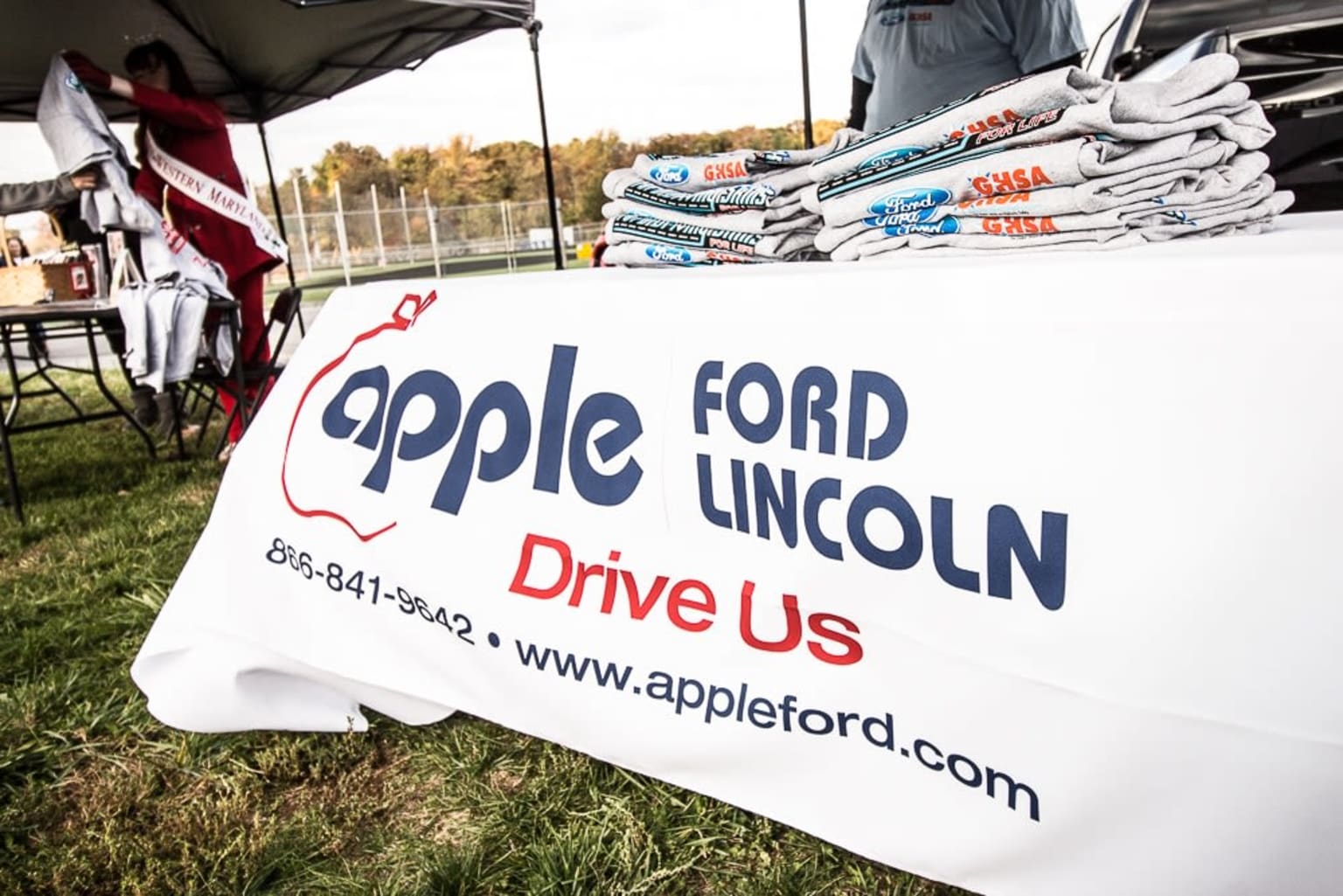 a table with a sign that reads apple ford lincoln drive us and a man standing next to a table with a pile of newspapers on it
