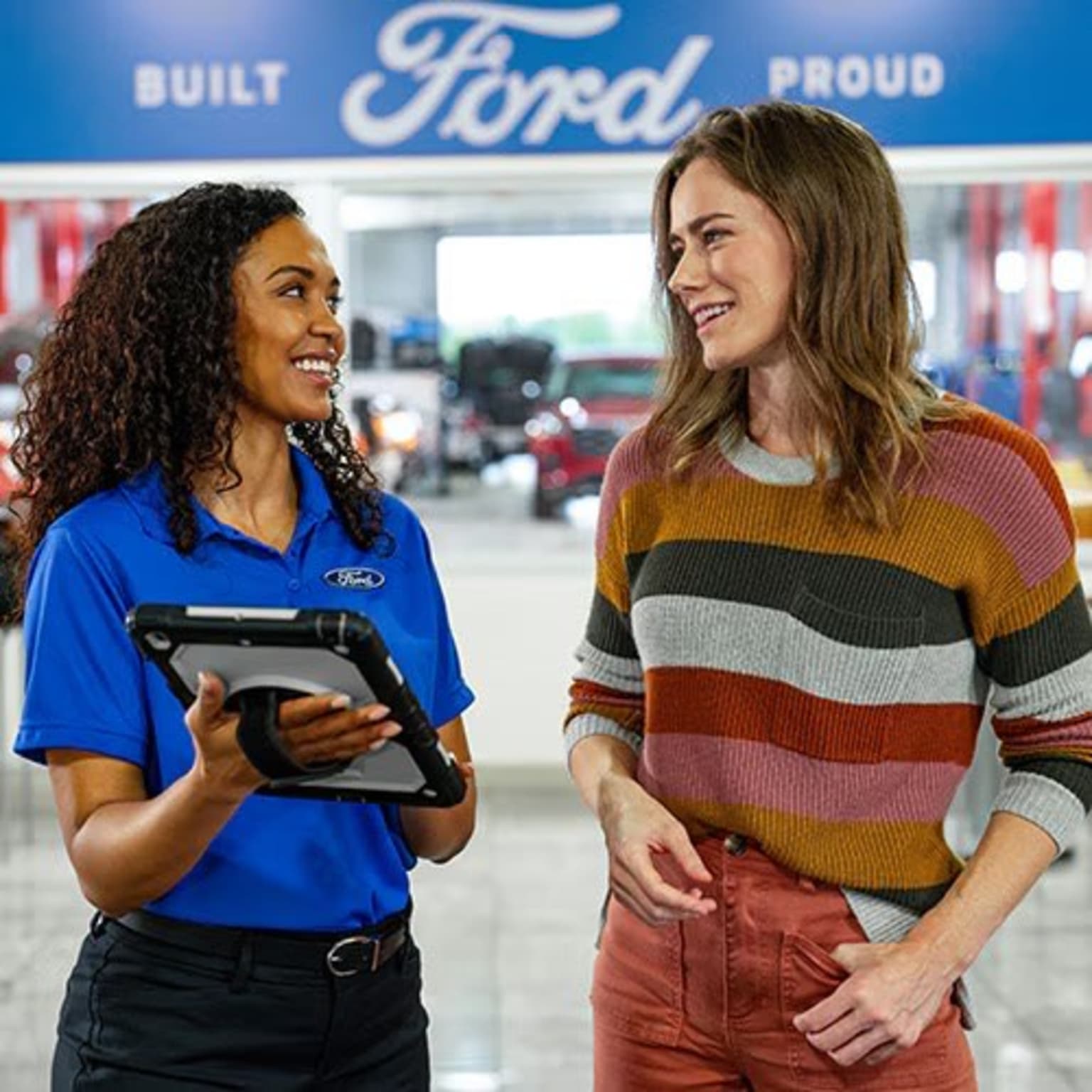 Two women, one wearing a blue uniform and the other a colorful striped sweater, are engaged in a friendly conversation in front of a dealership backdrop.