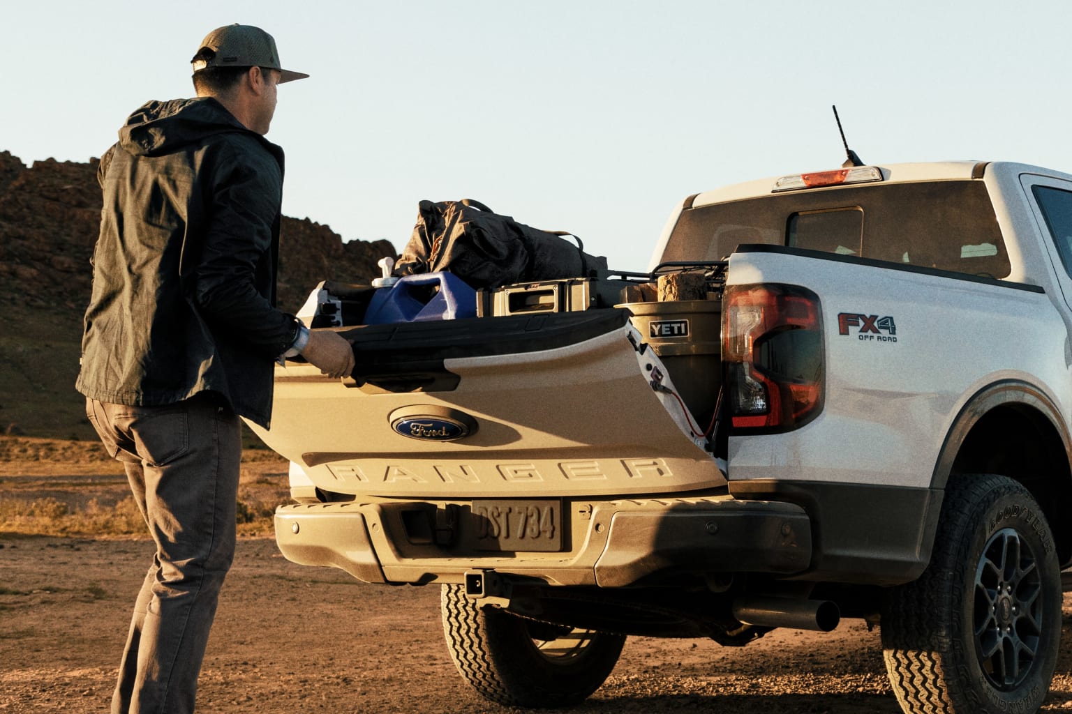 A person in a dark jacket and hat stands next to a white pickup truck in a desert landscape, loading gear and equipment into the truck's bed.