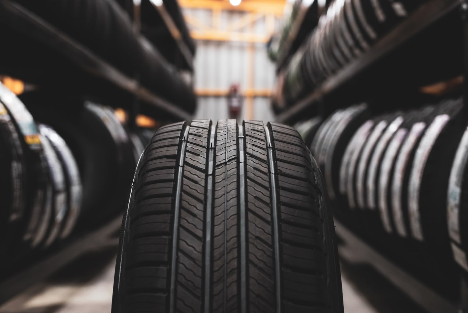 A single car tire stands in the foreground, centered between rows of stacked tires on shelves in a warehouse background.