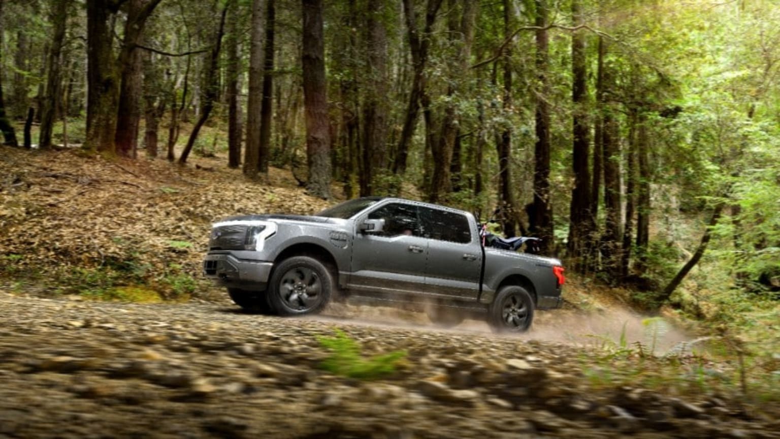 a silver truck driving down a dirt road in the middle of a forest with lots of trees on both sides of the road