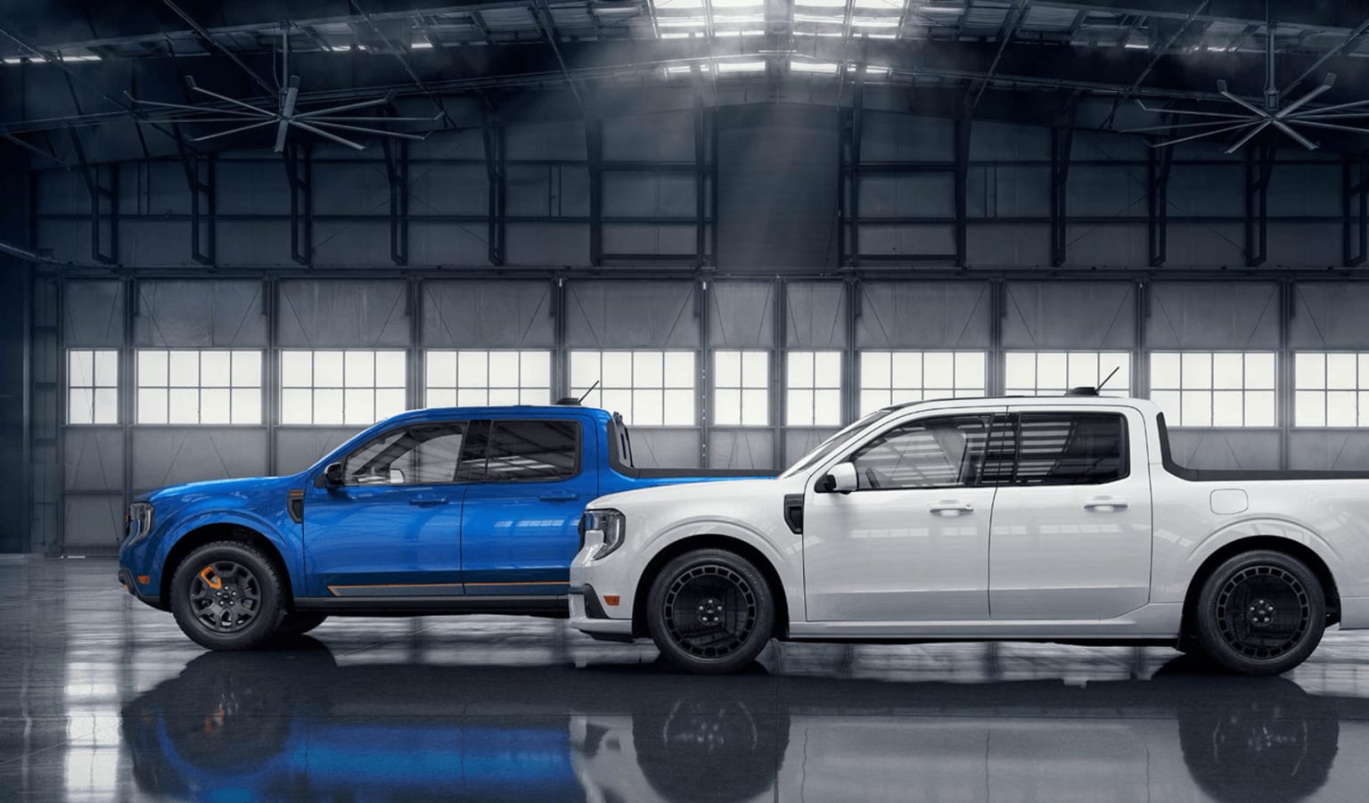 Two pickup trucks, one blue and one white, are parked in a large, dimly lit industrial warehouse with high ceilings and windows.