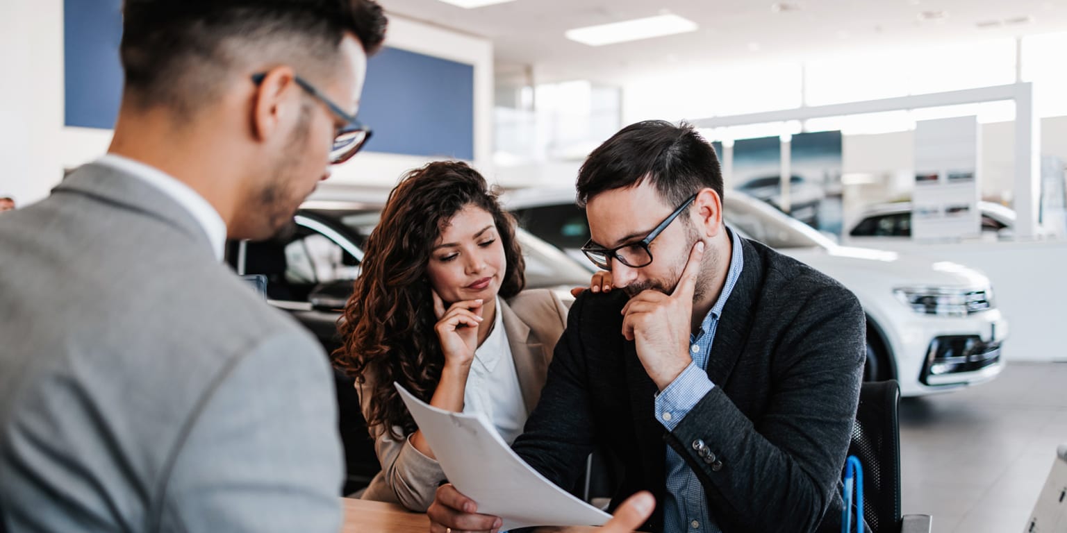 A salesperson in a gray suit discussing paperwork with a couple in a car dealership, with white cars displayed in the background.