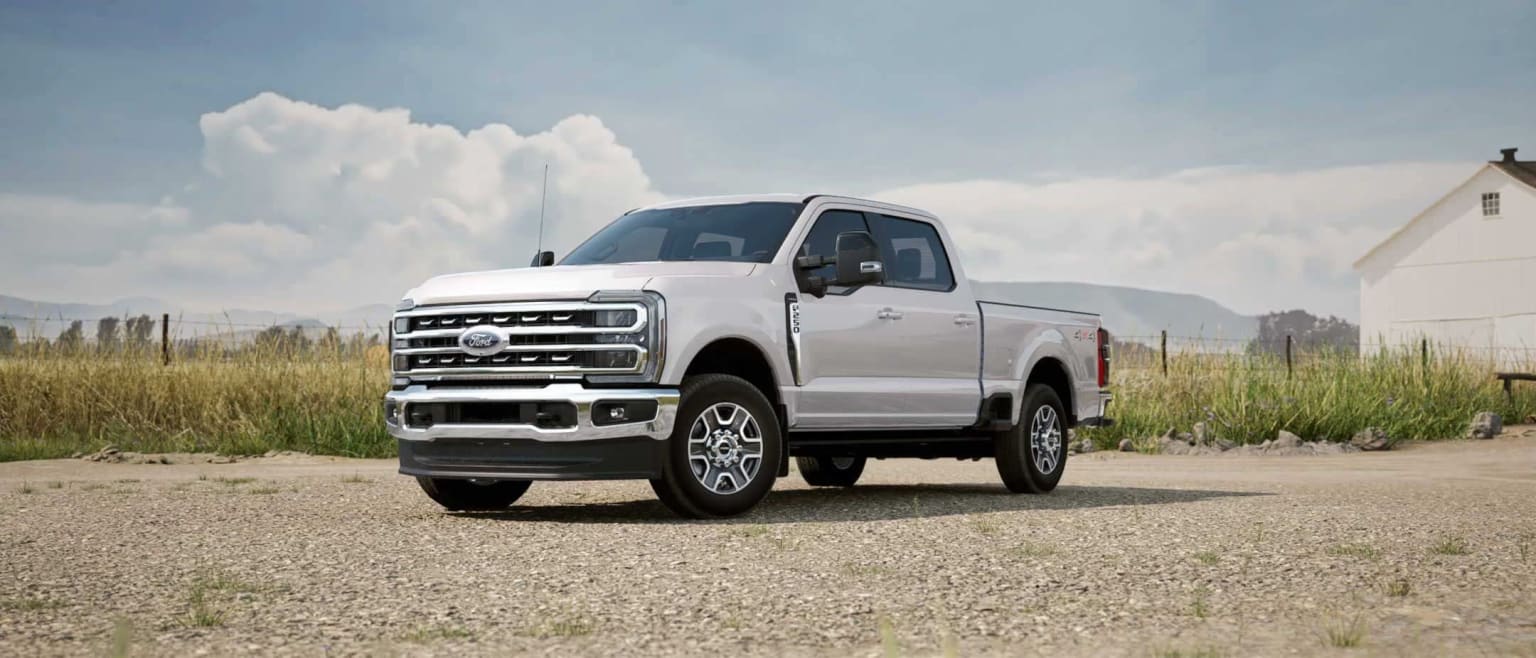 A white pickup truck on a dirt road with a grassy field and cloudy sky in the background.