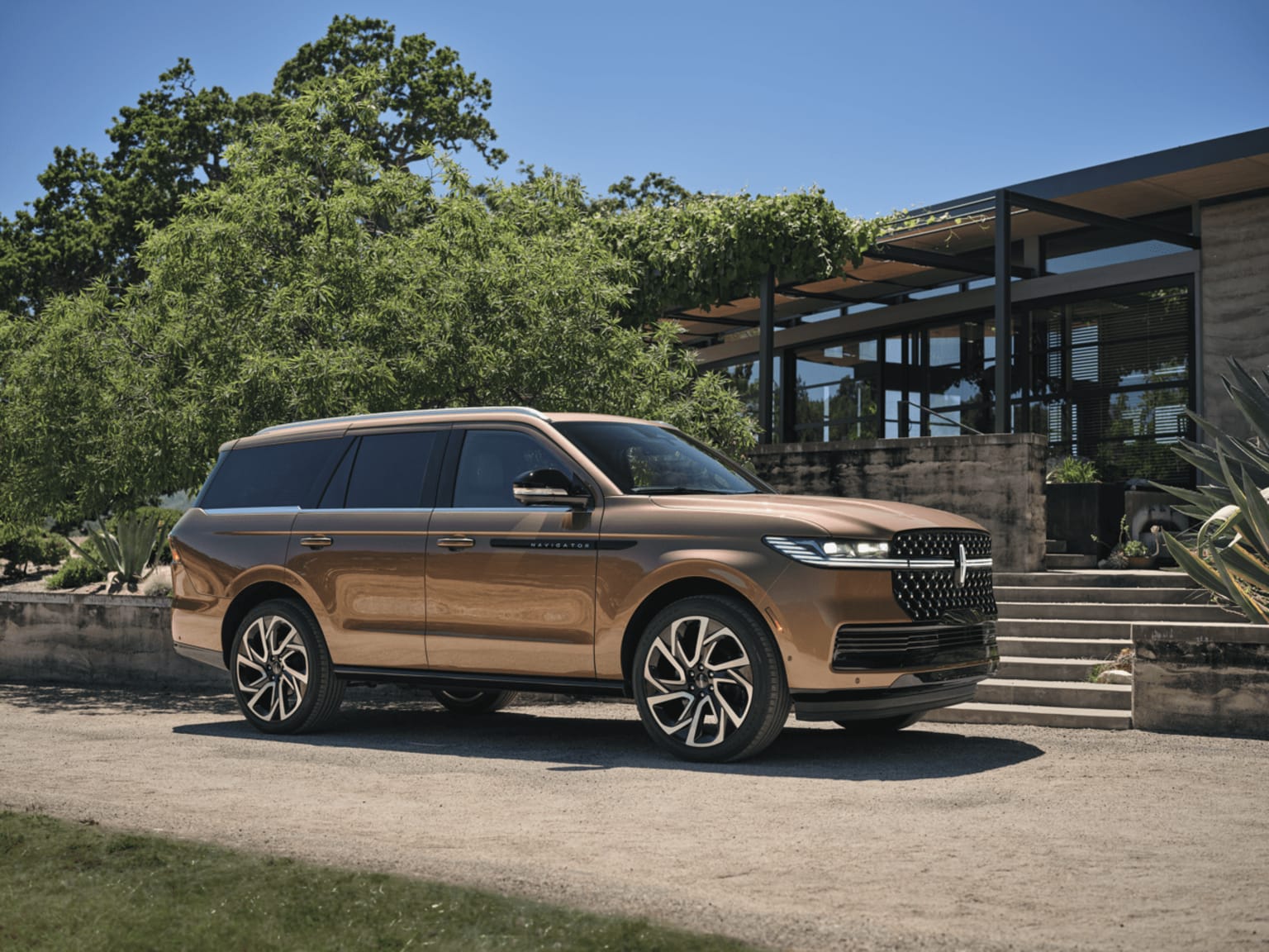 Large, luxurious SUV in a warm, metallic color parked on a gravel path in front of a modern, glass-walled building surrounded by lush, green trees.