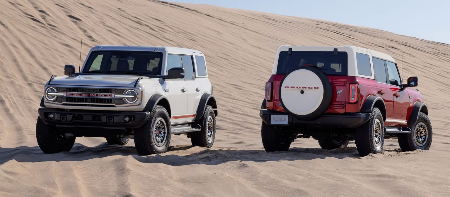 Two off-road vehicles parked in a desert landscape with sand dunes in the background.