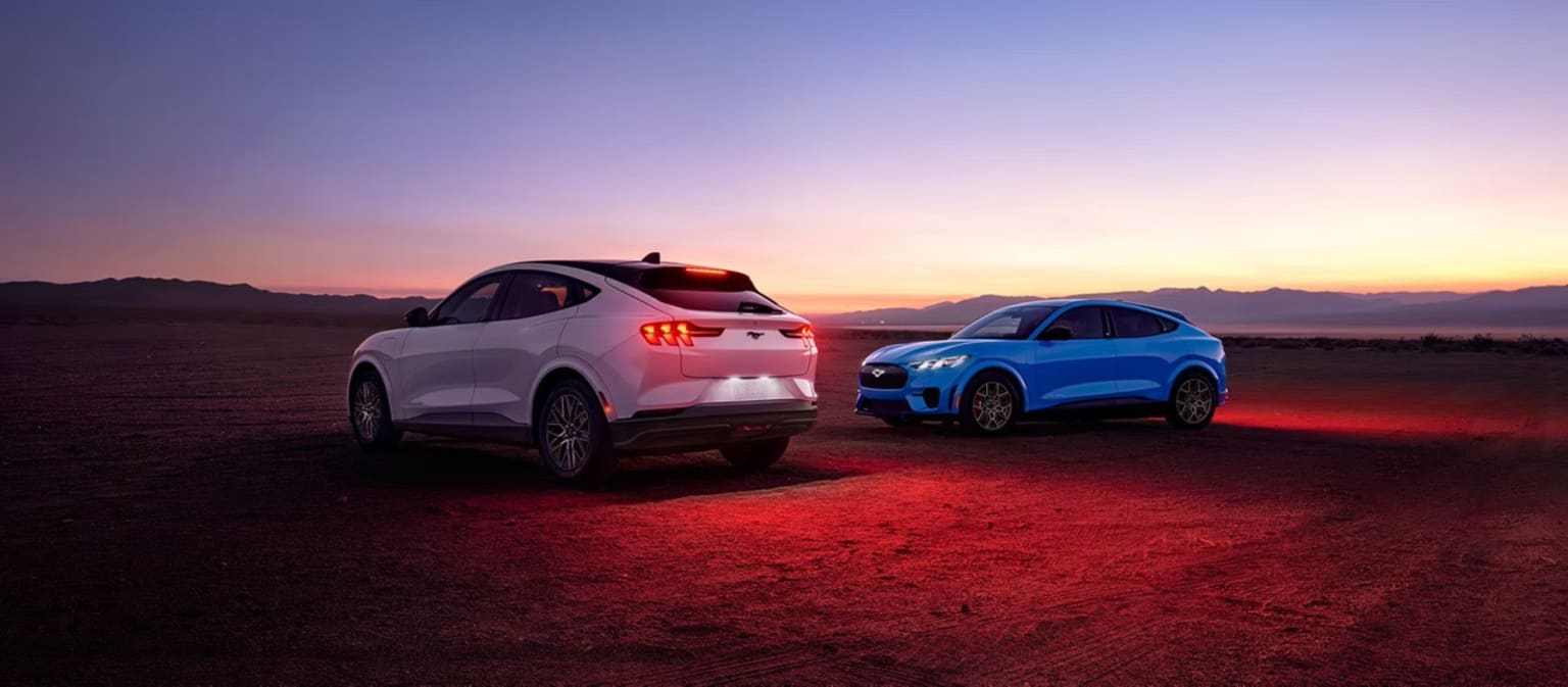 Two modern SUVs in a desert landscape with their headlights on, illuminating the rugged terrain at sunset.