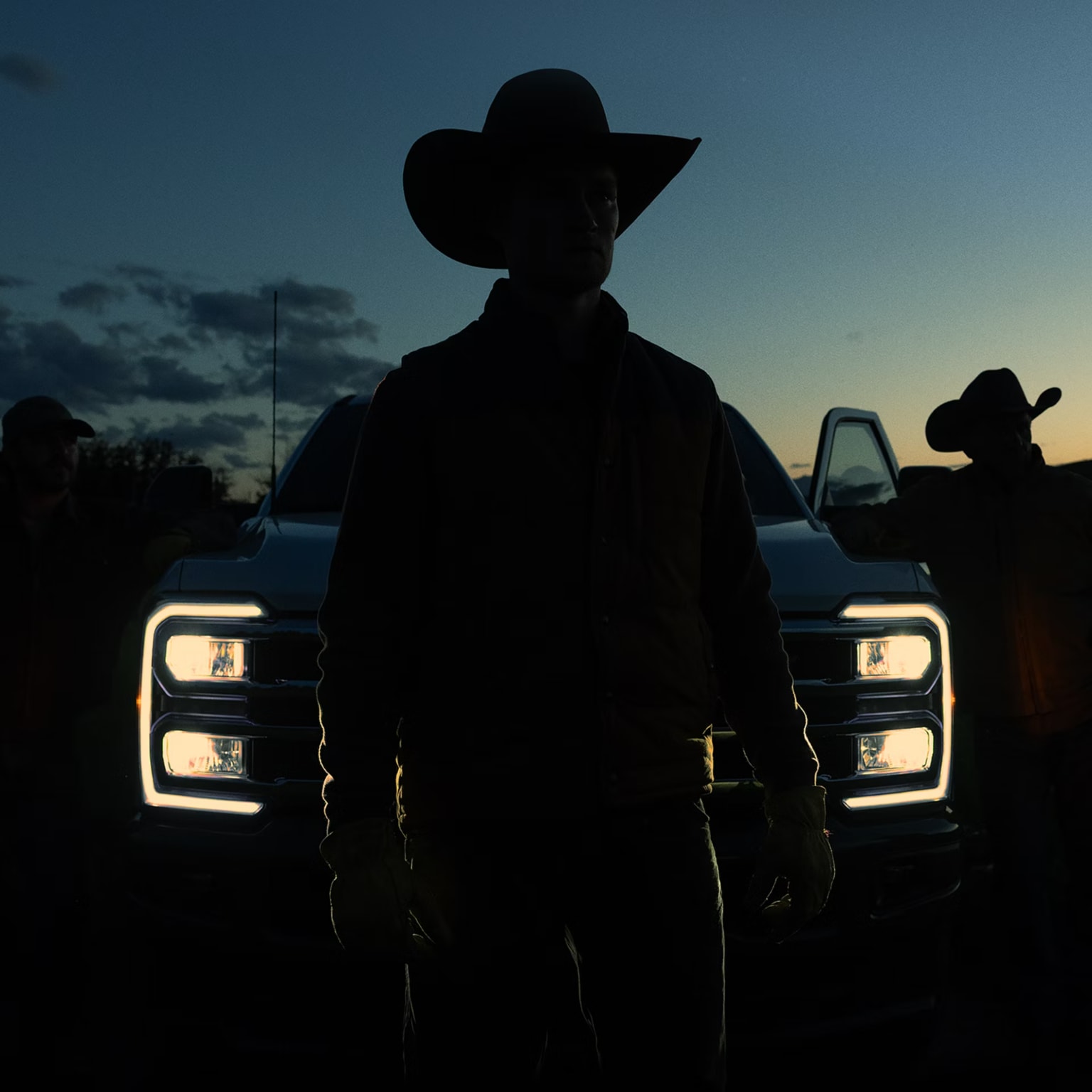 A silhouetted man in a cowboy hat stands in front of a white pickup truck with glowing headlights against a dark twilight sky.