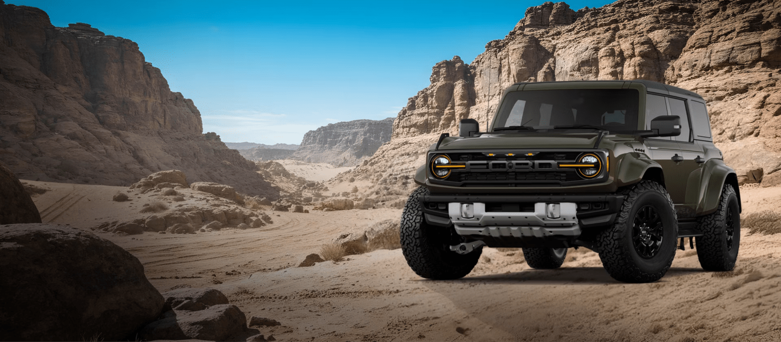 A dark green SUV is parked on a sandy desert floor with towering rock formations in the background under a clear blue sky.