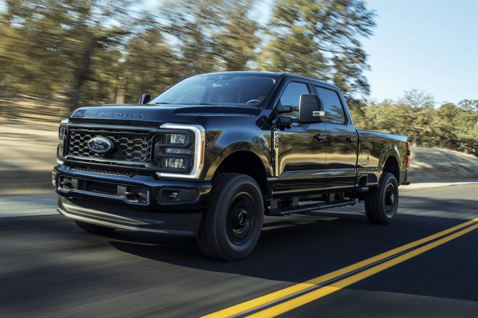 A powerful black Ford F-250 Super Duty pickup truck is speeding down a winding road surrounded by a lush forest landscape.