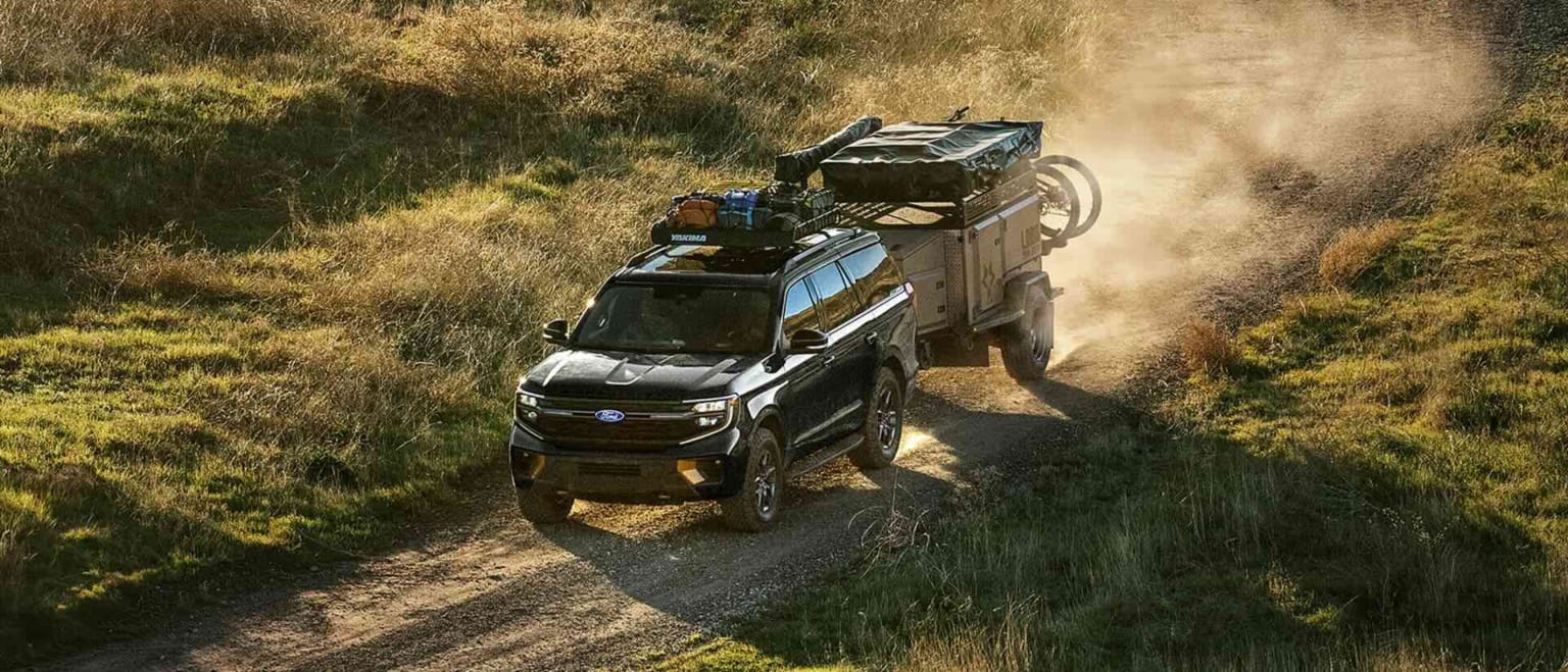 A black SUV towing a trailer along a dusty dirt road through a grassy, sunlit landscape.