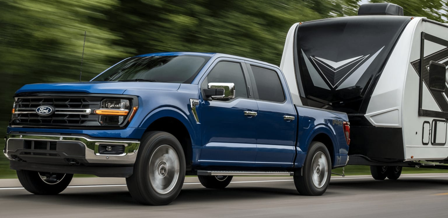 A blue pickup truck is towing a large white recreational vehicle against a blurred background of trees and greenery.