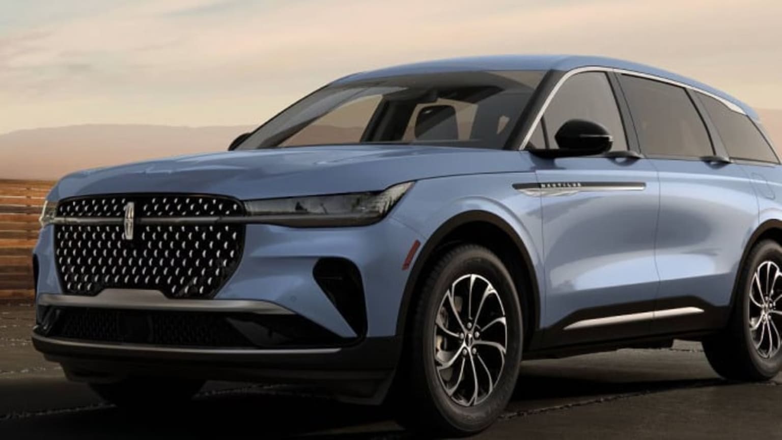 A light blue SUV with a distinctive grille parked on a dark road with a hazy, desert-like landscape in the background.