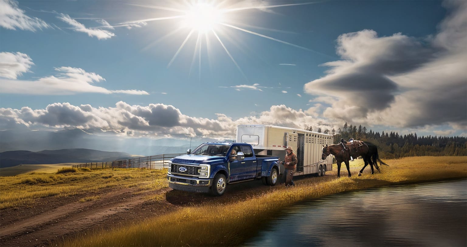Blue pickup truck with trailer parked on dirt road in scenic rural landscape, mountains and clouds in background, horse standing nearby.