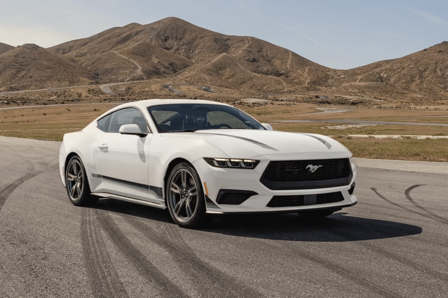 A white sports car is parked on a desert road, with rugged mountains in the background.