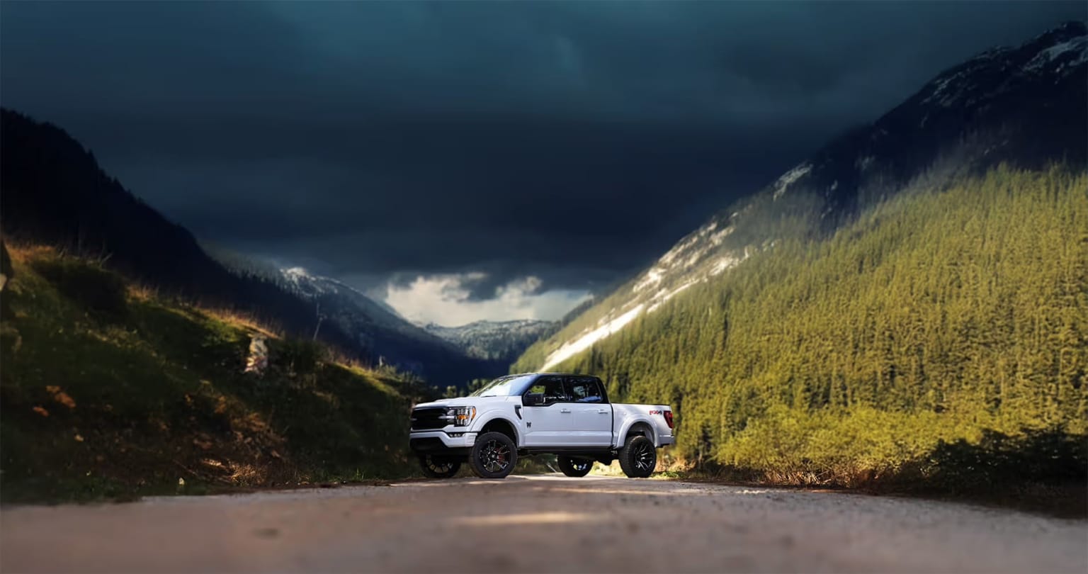 A white pickup truck on a winding mountain road with towering cliffs and lush, grassy slopes under a dramatic, stormy sky.