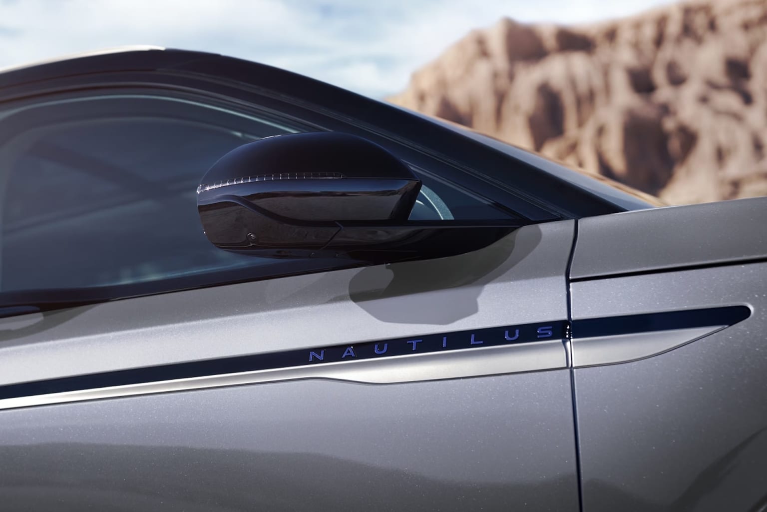A sleek, silver car with an unknown badge on the side is parked in front of a rugged, mountainous landscape.