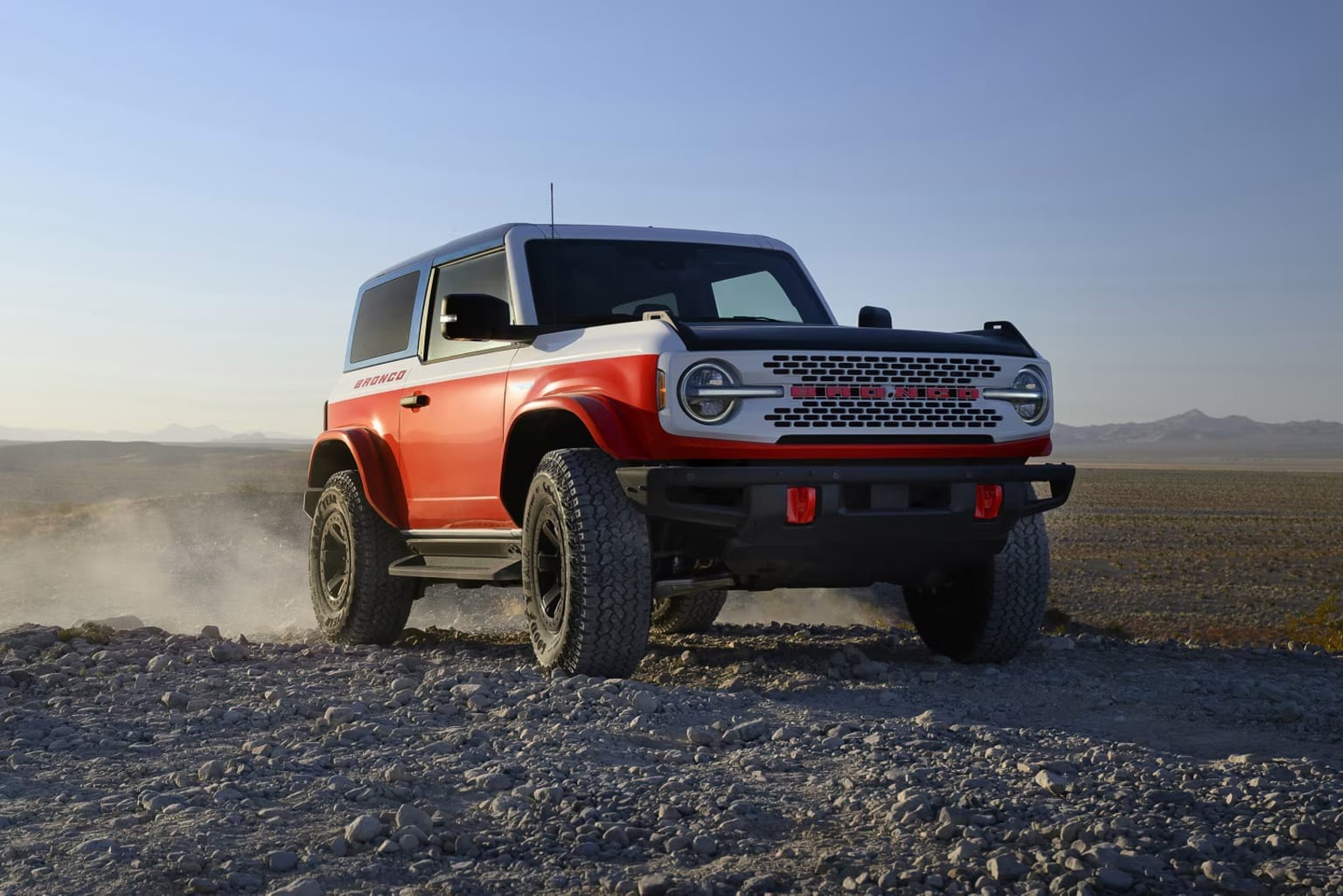 An off-road vehicle in a red and black color scheme driving through dusty, rocky terrain against a clear blue sky.