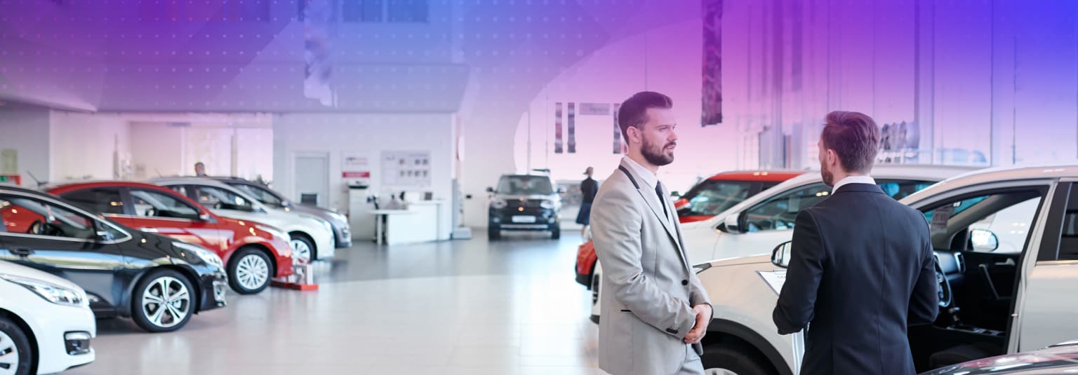 The image shows a car showroom with various vehicles on display, including red cars in the foreground. Two men, one in a suit and the other in casual attire, appear to be engaged in conversation in the middle of the showroom.