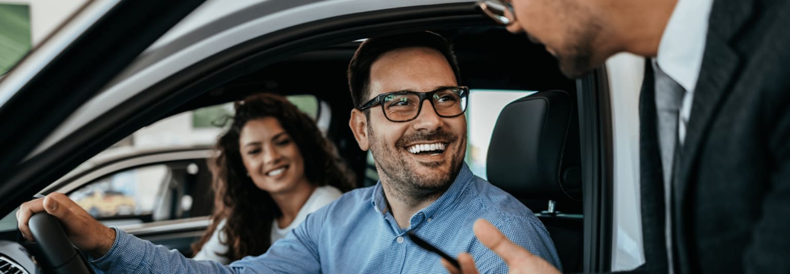A smiling man wearing glasses seated in the driver's seat of a car, with a woman sitting next to him in the passenger seat.